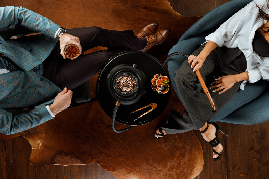 Overhead view of two people enjoying a luxury hookah session with Kaloud accessories, figs, and drinks on a dark wood table.