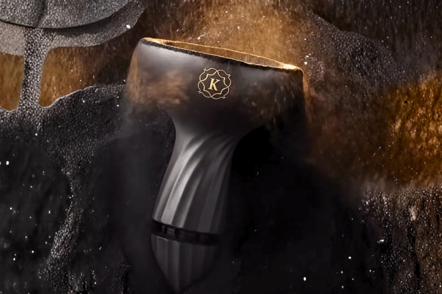 Luxury black hookah bowl head with gold rim and K logo surrounded by swirling tobacco smoke and particles on a dark premium background.