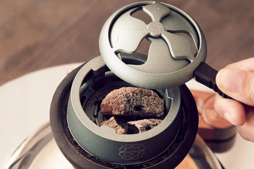 Kaloud Lotus heat management device with its vented lid lifted, showing lit charcoal held above the shisha in the hookah bowl